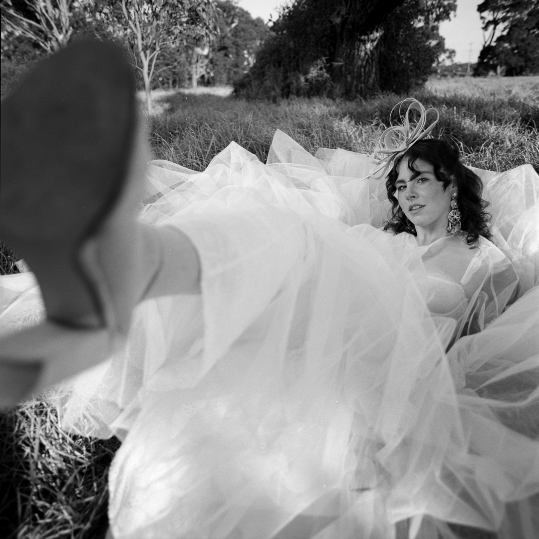 black and white film photograph of model wearing a tulle dress, sitting in a field kicking at the camera